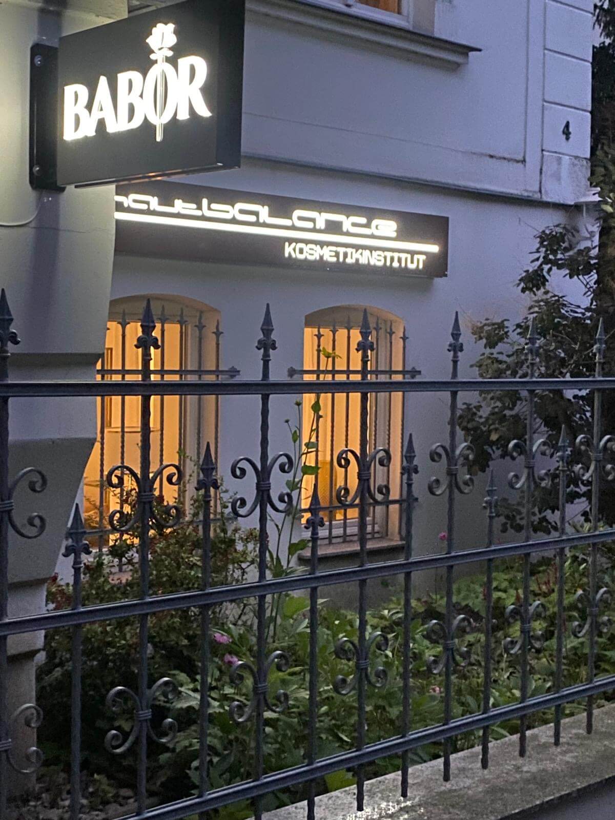 Ein beleuchtetes Schild eines Babor Kosmetikinstituts strahlt über einem gepflegten Gartenzaun.